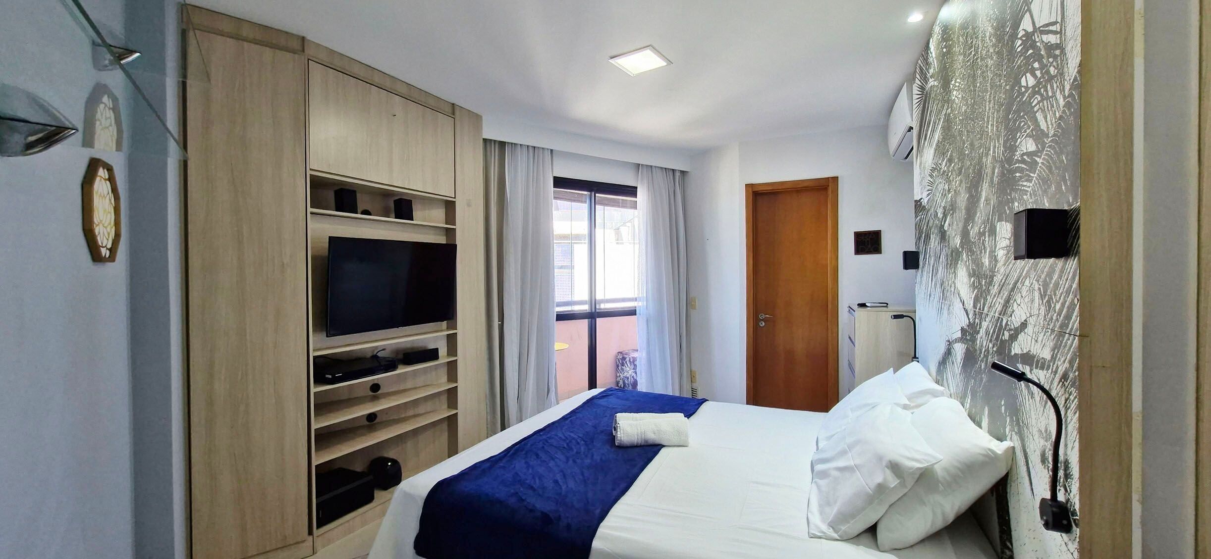 Ein Hotelzimmer mit Bett, Fernseher, Schrank, Fenster mit Vorhängen und einer Wand mit einem Blattmuster.