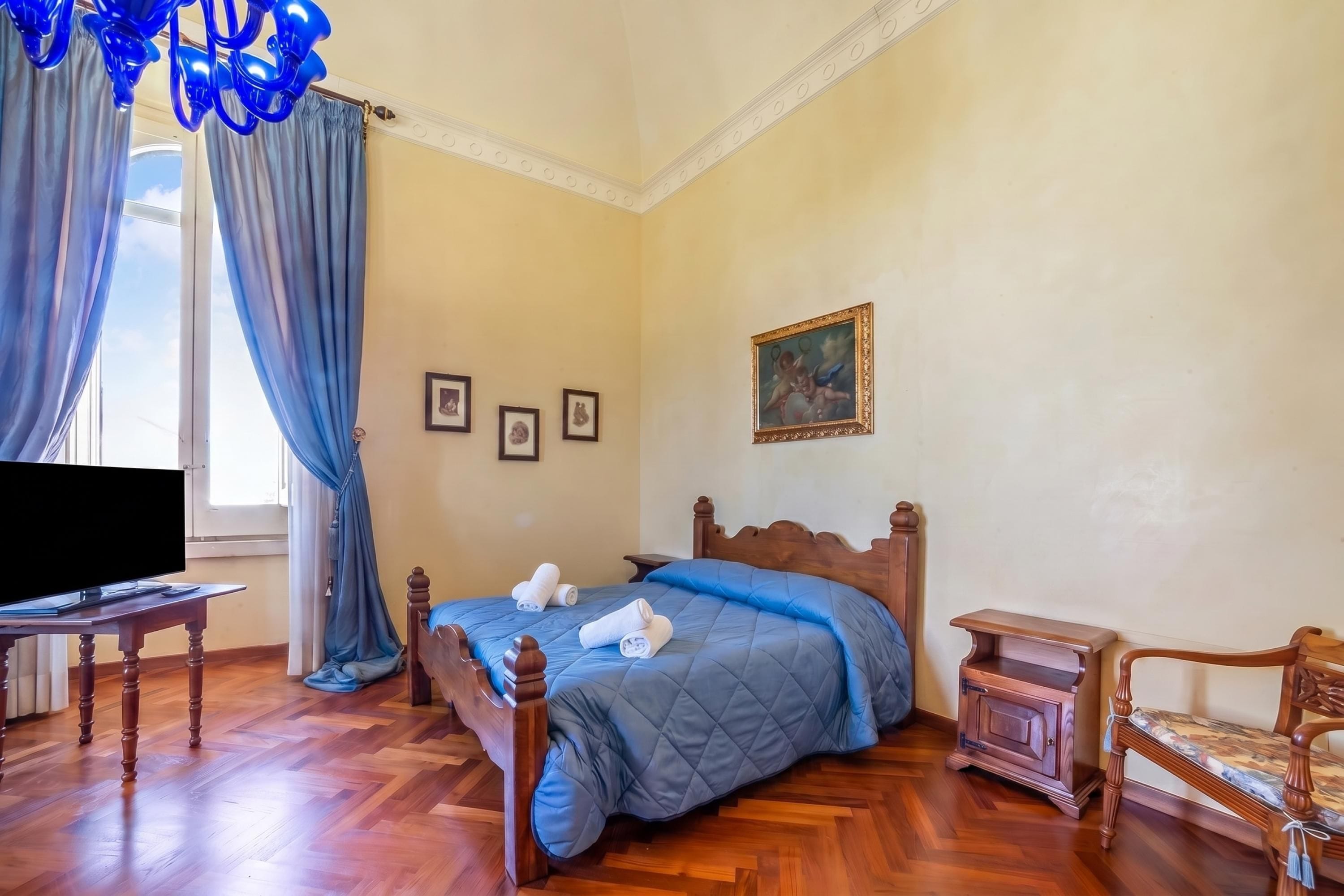 Un dormitorio con cama, televisor, mesita de noche de madera, una silla y un cuadro en la pared.