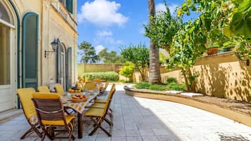 Un área de comedor al aire libre con una mesa y sillas, rodeada de follaje y un edificio con detalles arquitectónicos.