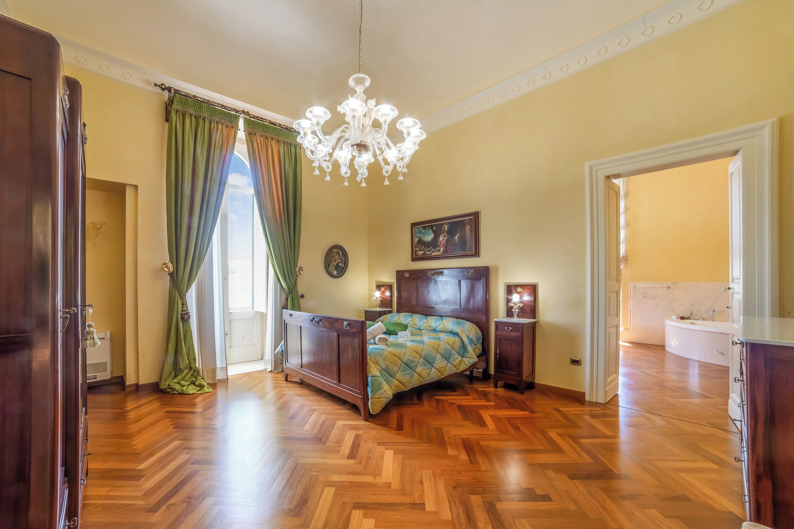 Quarto com piso de madeira, cama com colcha, lustre, quadro e banheiro visível através de uma porta aberta.