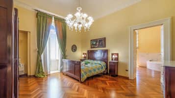 Quarto com piso de madeira, cama com colcha, lustre, quadro e banheiro visível através de uma porta aberta.