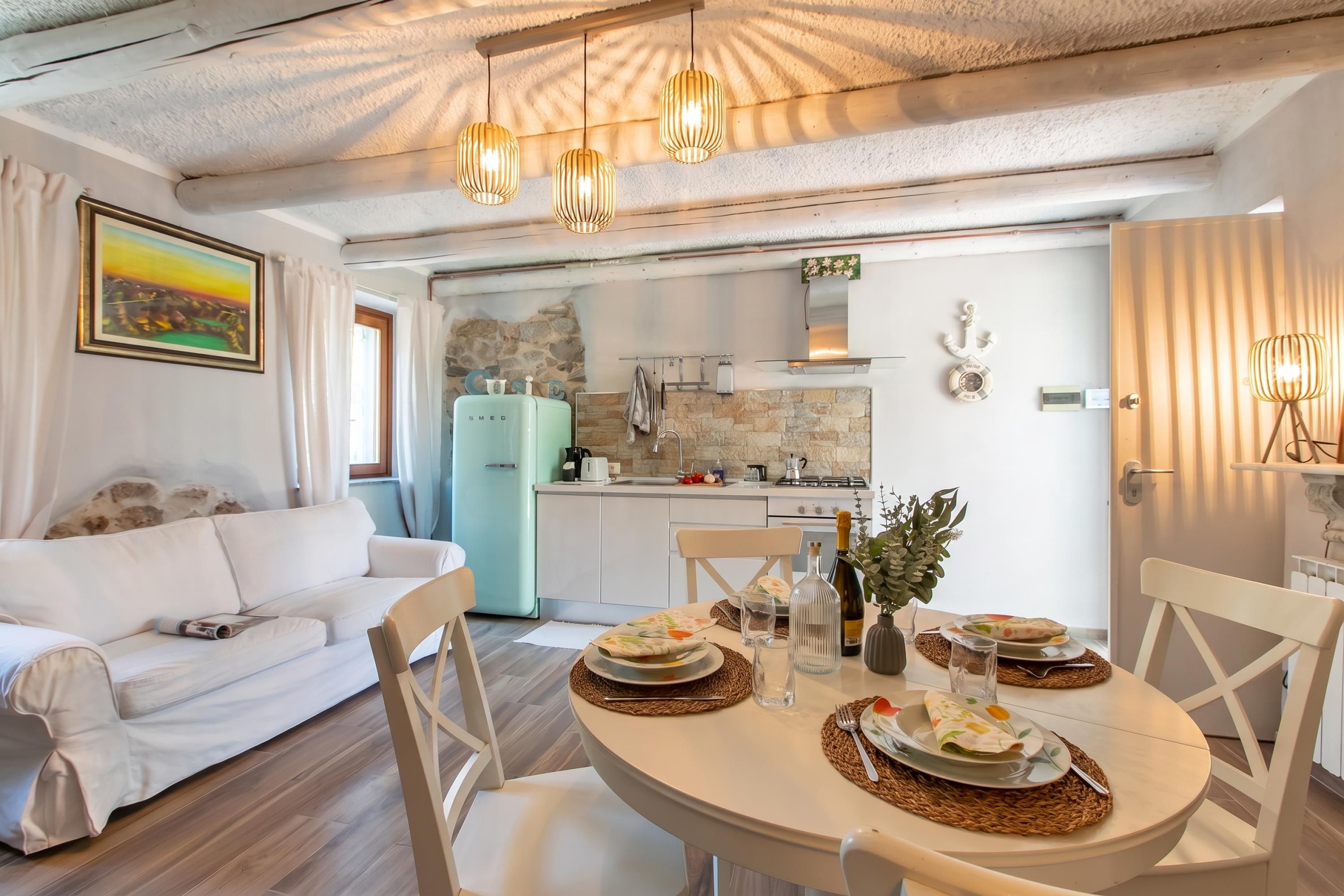 Un soggiorno moderno con un divano bianco, un tavolo da pranzo apparecchiato con piatti e bicchieri, e una cucina sullo sfondo con uno schienale in pietra.