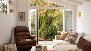 A living room with a brown leather chair, a white sofa with patterned cushions, and a large sliding glass door leading to a garden. - Wondervale - Escape to the Southern Highlands (Burrawang)