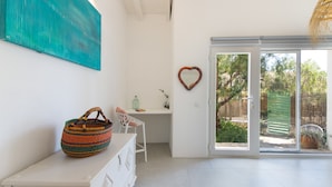 Una habitación con un escritorio blanco, una silla, una cesta grande de fibra natural, un espejo con forma de corazón y un ventanal corredero que da a un espacio al aire libre.