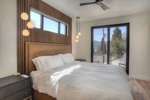Ein Schlafzimmer mit einem großen Bett, einem Holz-Kopfteil, einem Nachttisch und einer Sitzgelegenheit am Bett, mit Blick ins Freie durch eine Schiebetür.