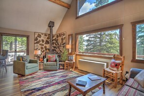 A cozy living room with a stone wall, a fireplace, and large windows offering a view of trees. - New Listing! Quiet Mtn Cabin | River and Mtn Views | Expansive Deck (Creede)
