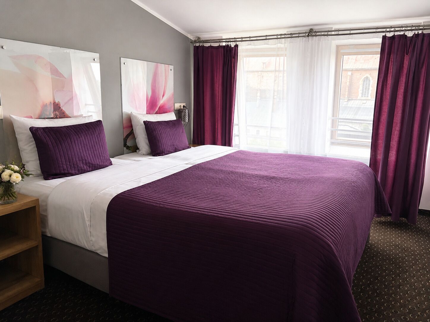 A hotel room with a large bed, two pillows, a nightstand with a vase of flowers, and a window with curtains.