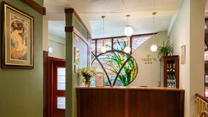 A reception area with a checkered floor, a reception desk, a wall-mounted clock, a framed artwork, a stained glass window, and a sign with a logo and stars. - Hotel Secesja (Kraków)