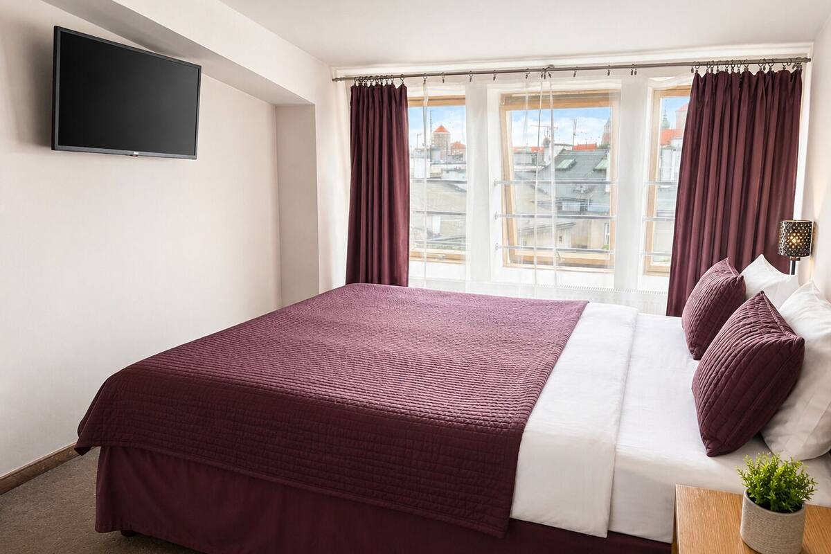 A hotel room with a large bed, a flat-screen TV mounted on the wall, and a window with curtains.