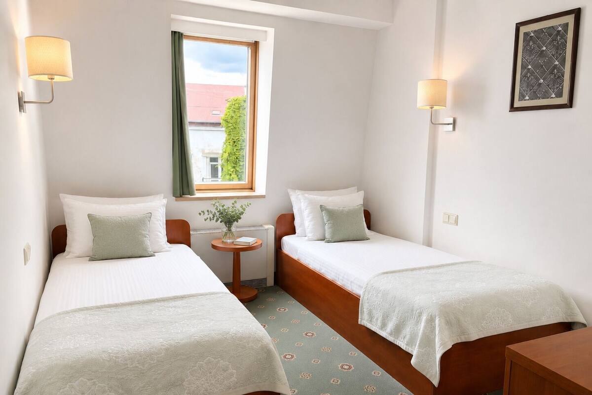 A small, clean room with two beds, a window with curtains, a small table with a vase, and a framed picture on the wall.