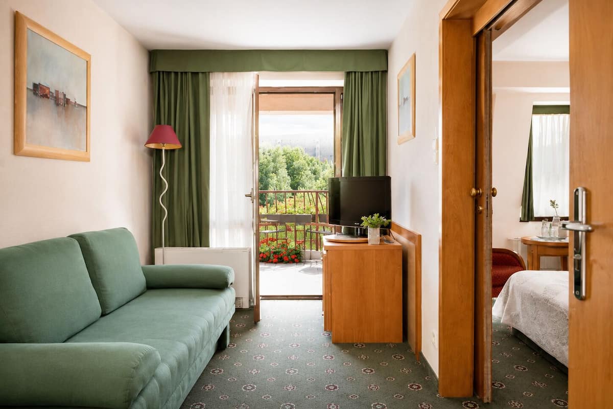 A hotel room with a green sofa, a television on a wooden stand, a balcony with a view of greenery, and a door leading to another room.