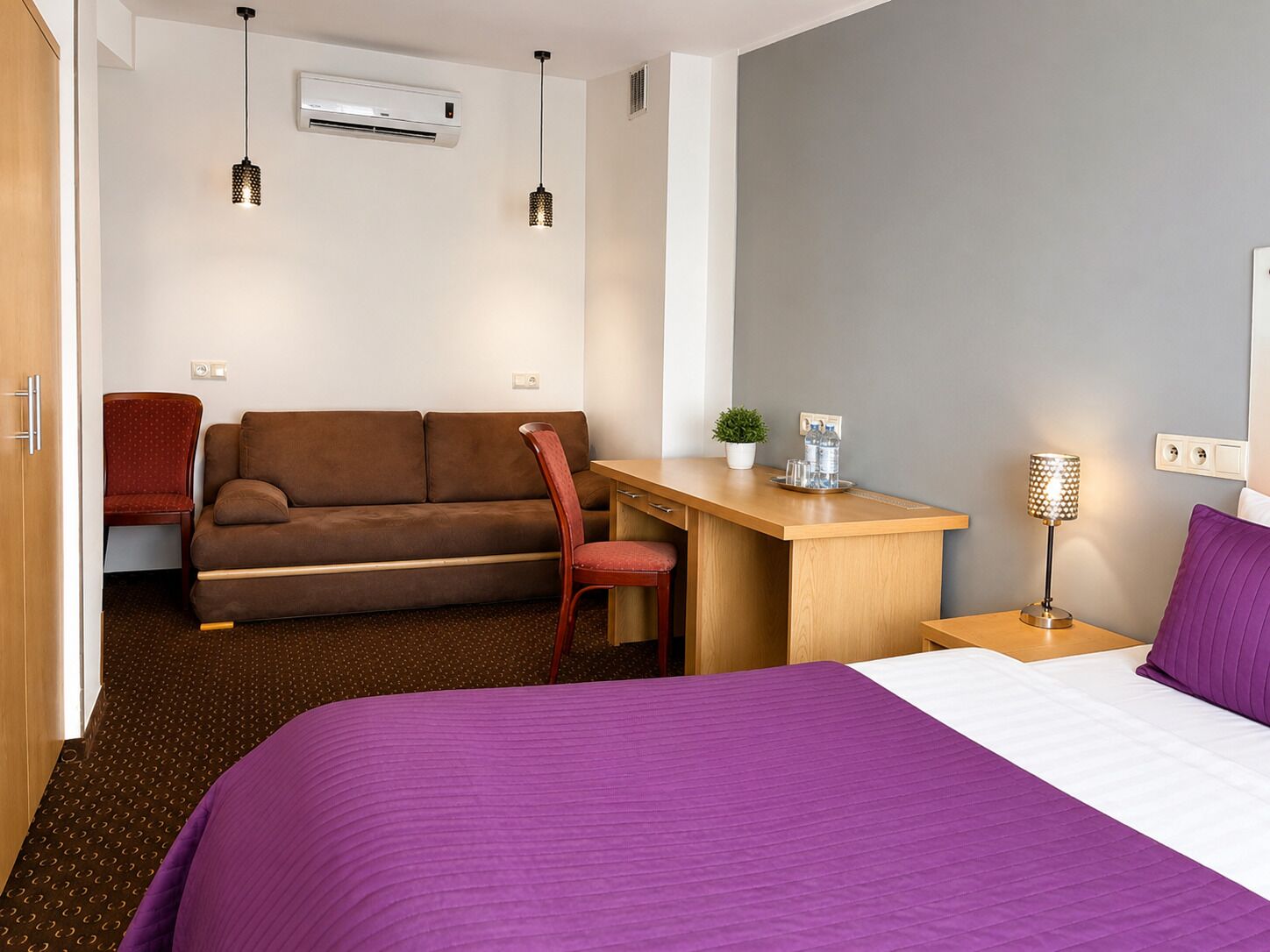 A hotel room with a bed, a desk, a sofa, a chair, an air conditioner, and a lamp.