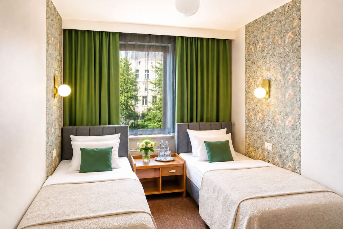 A hotel room with two beds, green curtains, a window with a city view, and a small table with a vase.
