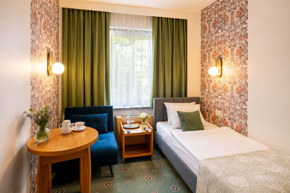 A hotel room with a bed, a desk, a chair, a small table with a cup and saucer, and a vase with flowers.
