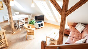 Ein Loft-Zimmer mit Holzbalkendecke, einem Fernseher, einer Couch und einer Essecke mit Stühlen und Tisch.