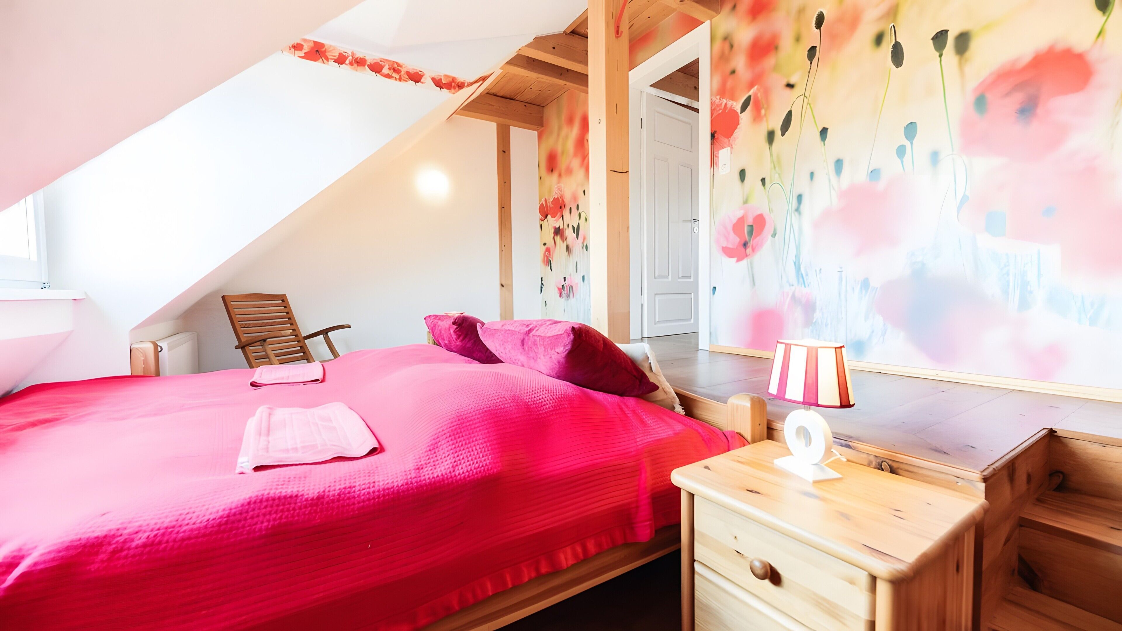 A bedroom with a sloped ceiling, a bed with a red bedspread, a wooden chair, a bedside table with a lamp, and a wall with a floral mural.