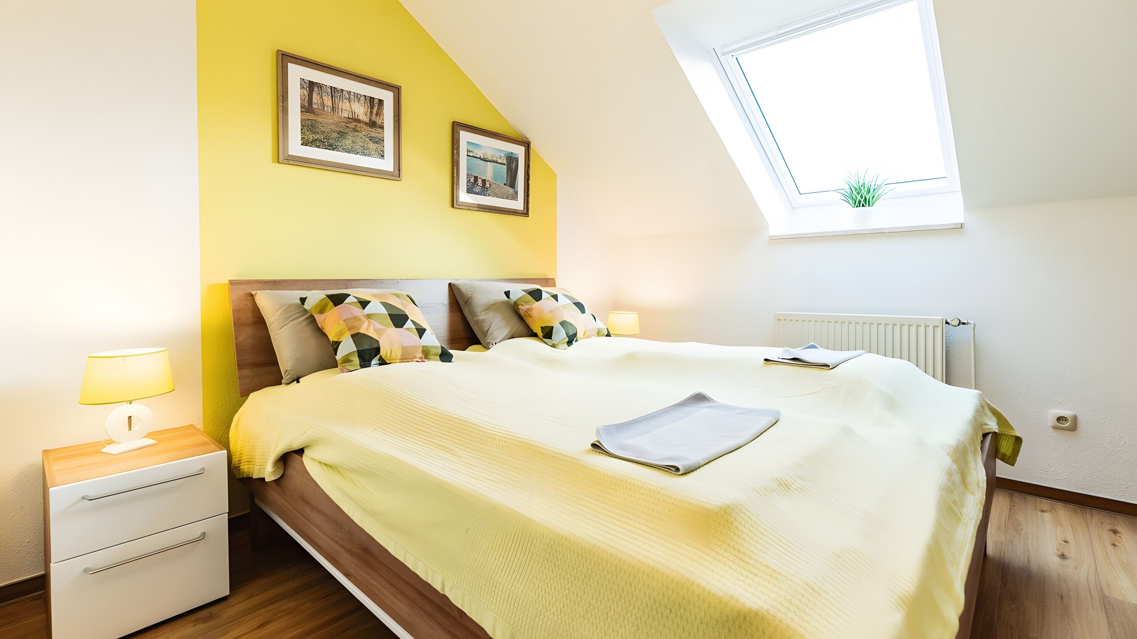 A bedroom with a bed, a nightstand, a lamp, a window with a plant, and two framed pictures on the wall.