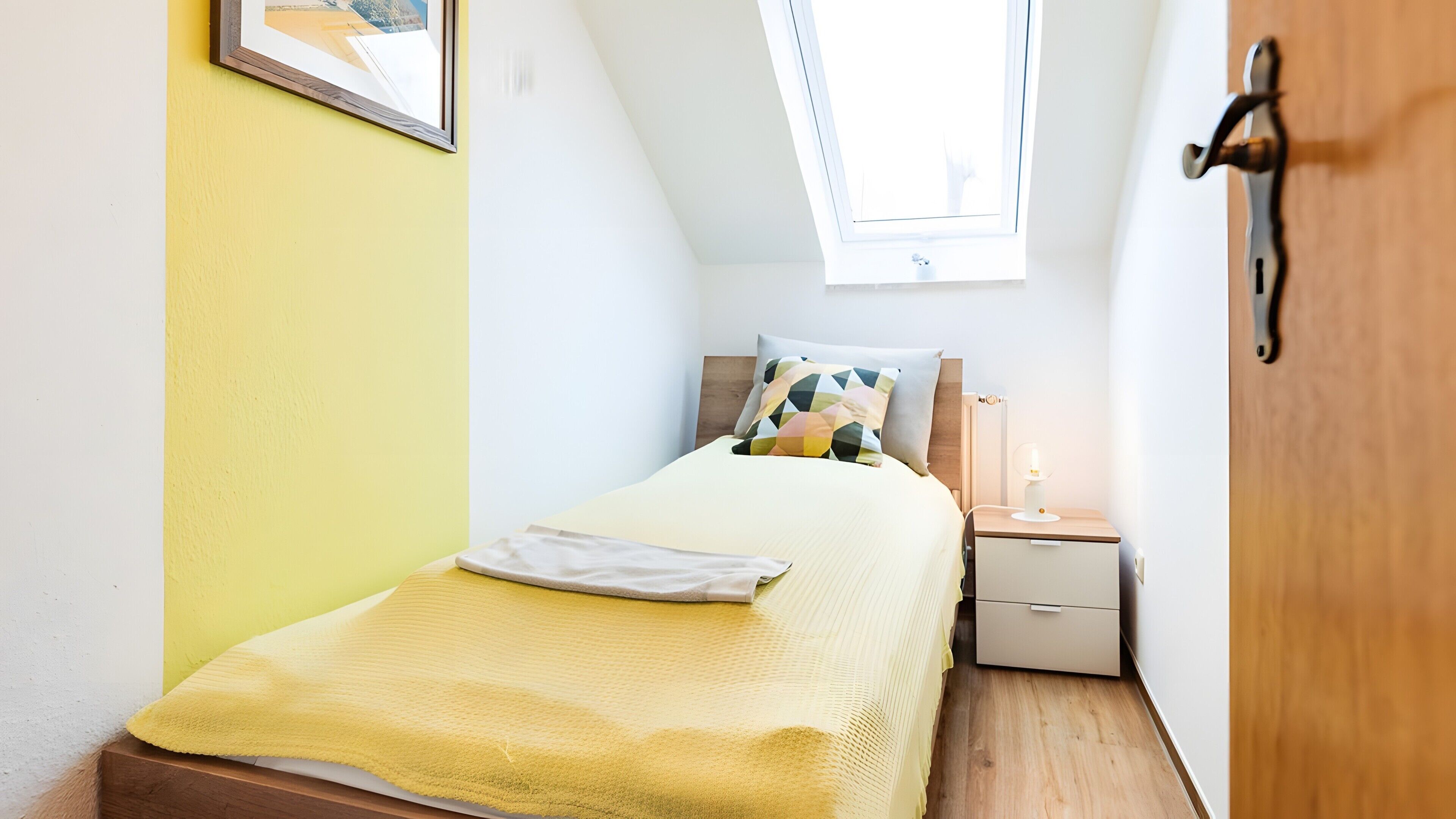 Un dormitorio pequeño y bien iluminado con cama, mesita de noche, lámpara y ventana.