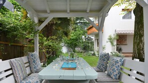 A covered patio with a table set for dining, surrounded by white benches with patterned cushions. - Double Room with Bathroom and Balcony, Mountain View (Merano)