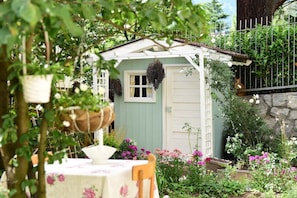 Ein charmanten Garten mit einem kleinen Schuppen, einem Tisch mit einer blumigen Tischdecke sowie verschiedenen Pflanzen und Blumen.