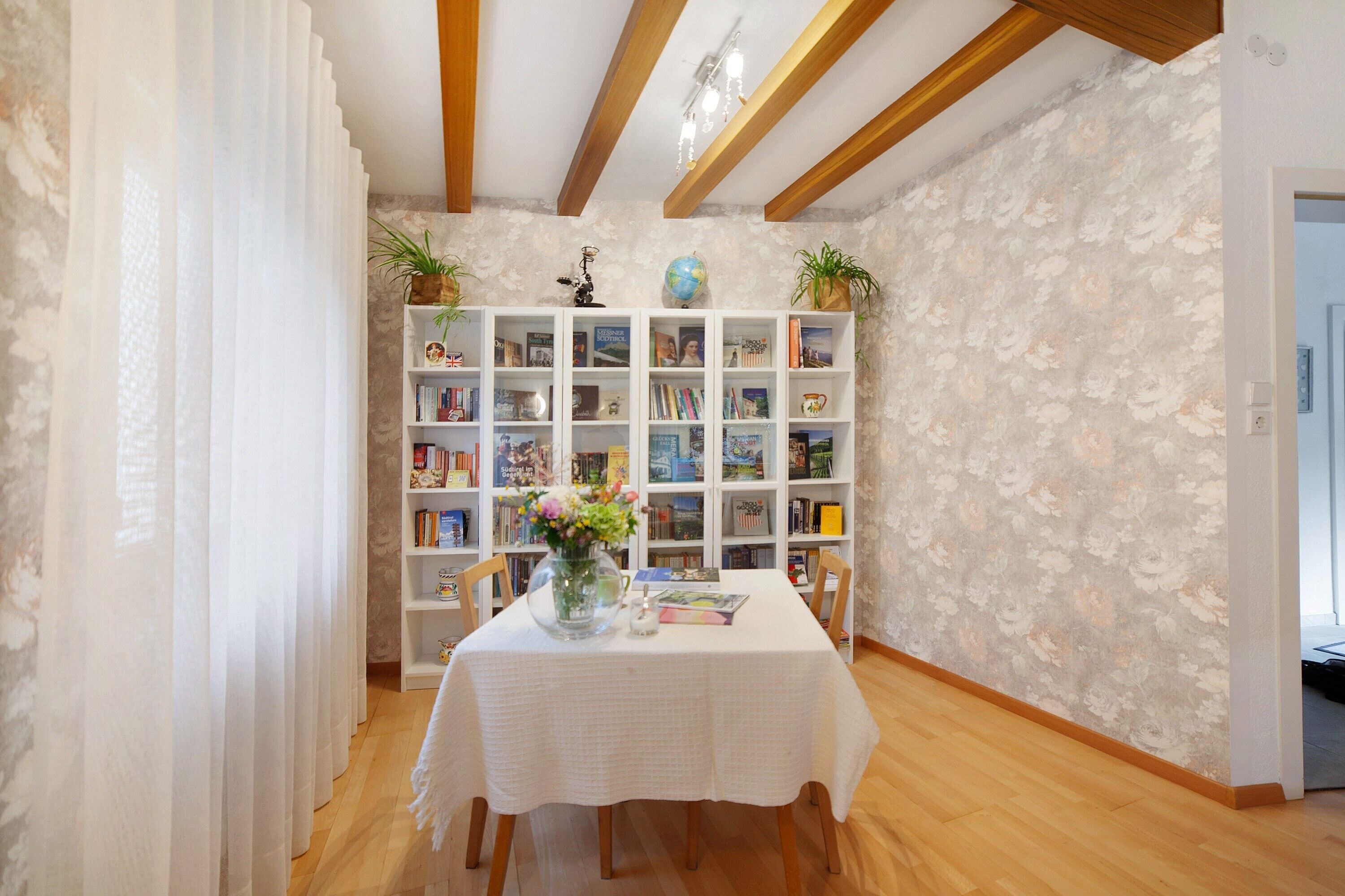 A room with a wooden floor, white walls, a white table with a tablecloth, and a bookshelf filled with books and decorative items.