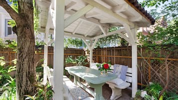 A covered patio with a table and bench, surrounded by greenery and a wooden fence.