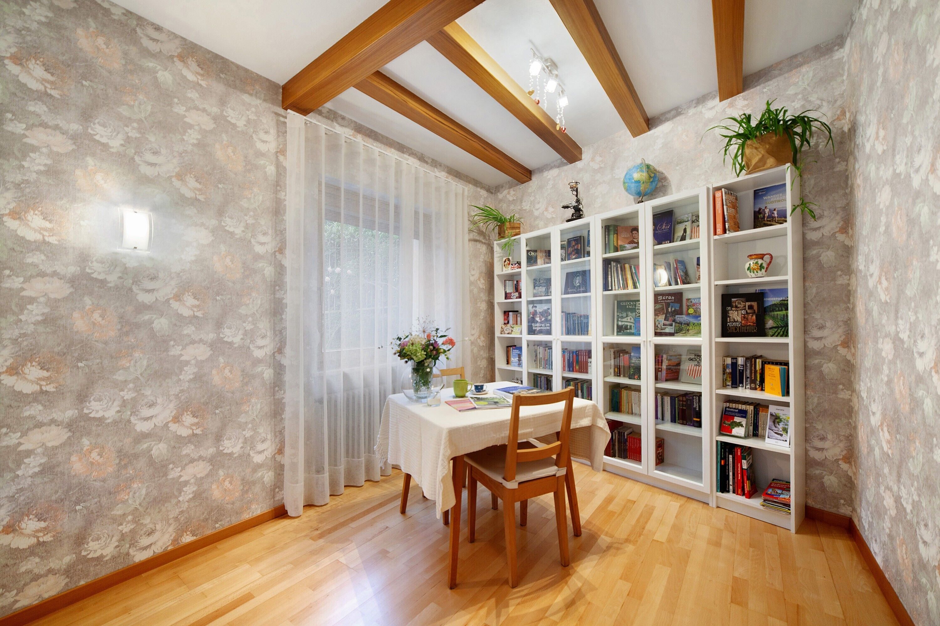 A dining area with a table, chairs, a bookshelf, and a window with sheer curtains.
