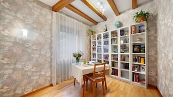 A dining area with a table, chairs, a bookshelf, and a window with sheer curtains.