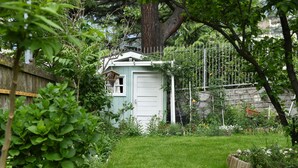 Un petit abri de jardin avec une porte blanche, entouré d’un jardin où poussent différentes plantes et délimité par une clôture en bois.