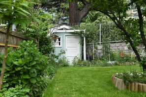 Un cobertizo pequeño con una puerta blanca, rodeado por un jardín con diversas plantas y una valla de madera.