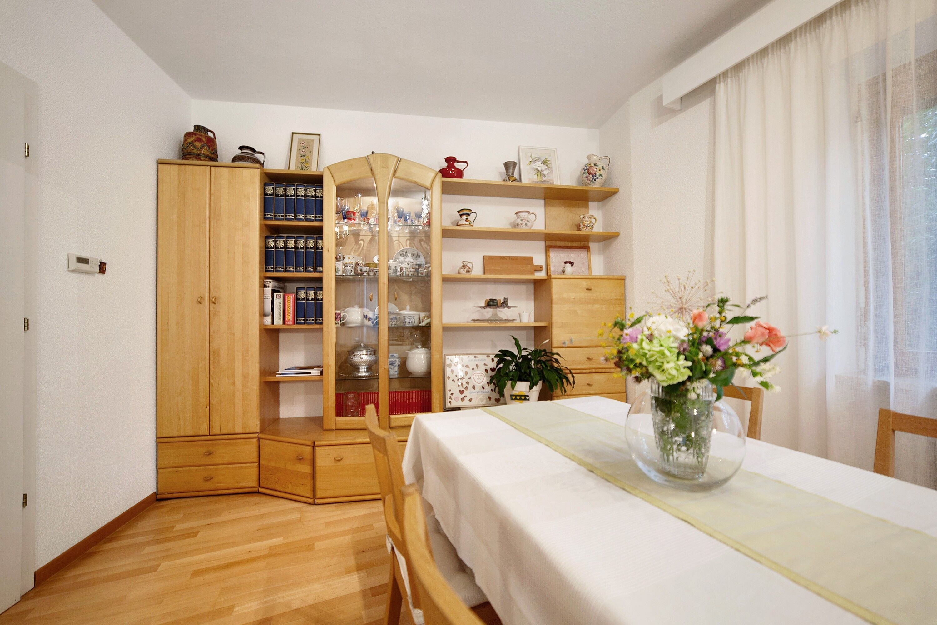 Une pièce avec un meuble en bois, une table recouverte d’une nappe blanche et des étagères remplies de divers objets.