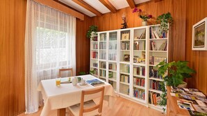 Une pièce chaleureuse aux murs en bois, ornée d’un rideau en dentelle, avec une bibliothèque blanche et une table sur laquelle se trouvent un livre et un verre de jus de fruits.