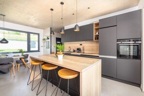 Eine moderne Küche mit einer zentralen Kochinsel, Barhocker, Pendelleuchten und Blick durch große Fenster.