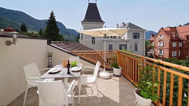 A balcony with a table set for two, white wicker chairs, a potted plant, and a view of traditional buildings and mountains. - Villa Thaler Apartment with Mountain View and Rooftop Terrace (Merano)