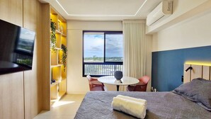 A modern hotel room with a bed, a TV, a small table with chairs, a balcony with a view, and a bookshelf. - COSTE0112 - Premium Stella Maris (Salvador)