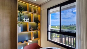 A room with a large window, a wooden shelf with decorative items, and a red chair. - COSTE0112 - Premium Stella Maris (Salvador)