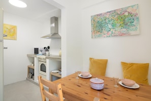 Una cocina compacta con electrodomésticos blancos, una mesa de comedor con cojines amarillos y un cuadro abstracto de colores en la pared.
