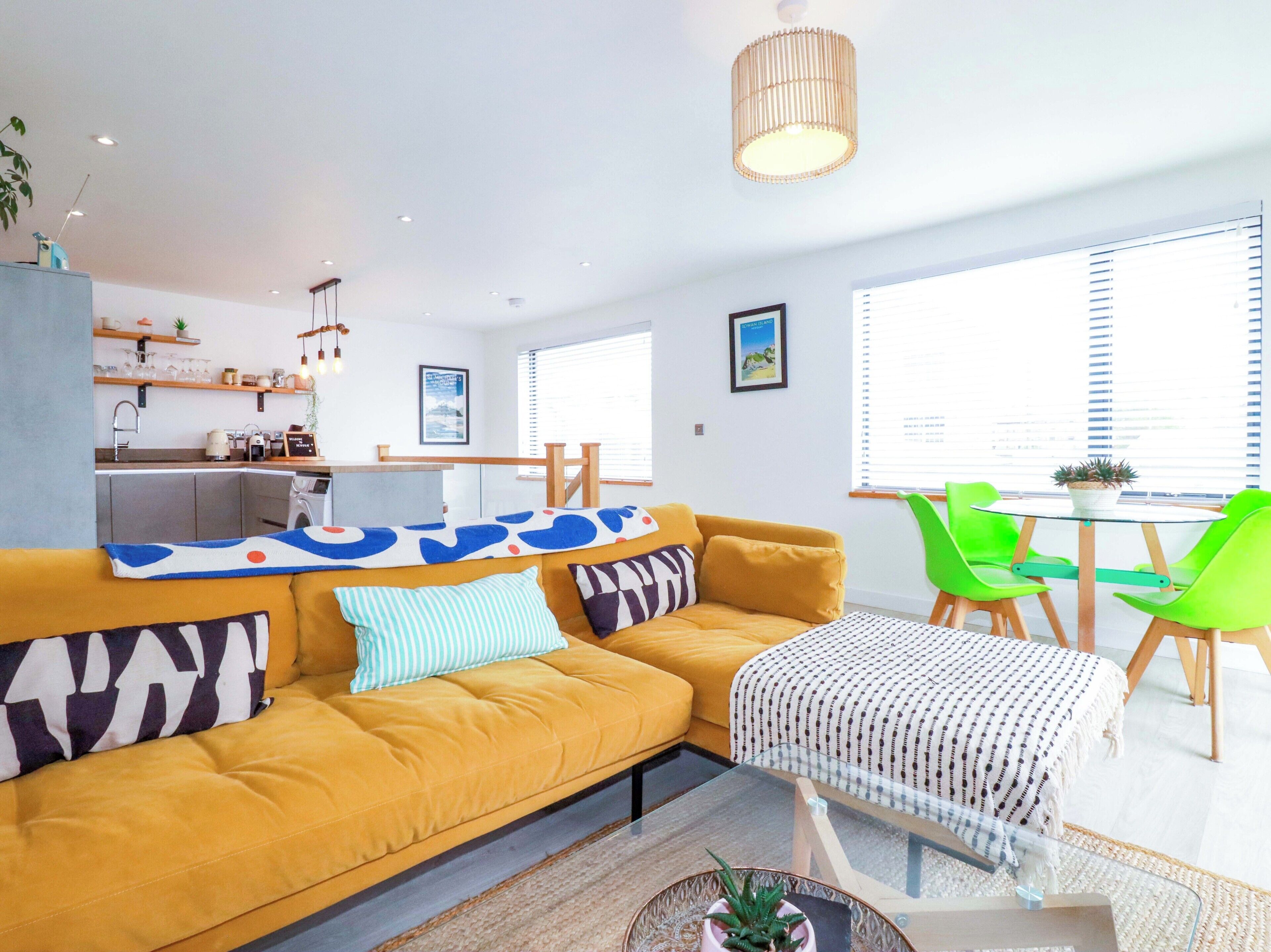 A modern living room with a yellow sofa, a glass coffee table, green chairs, and a kitchen area in the background.