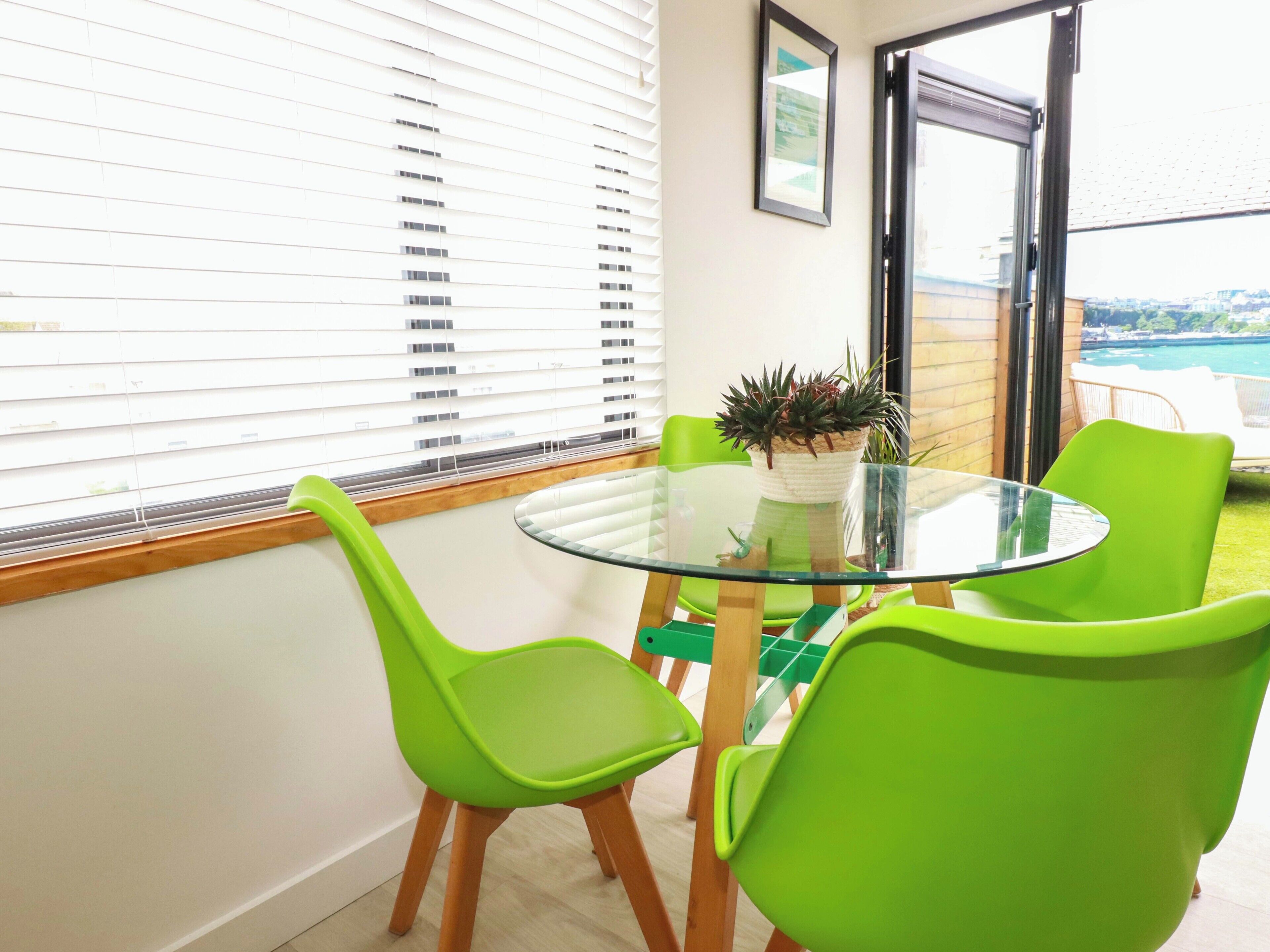 A room with a glass table, green chairs, a potted plant, and a view through a window.