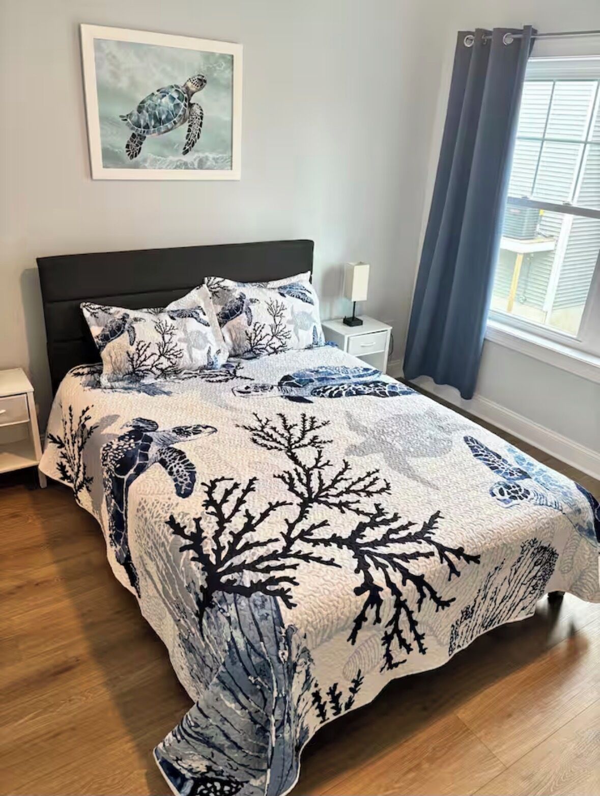 Une chambre à coucher avec un lit, une table de chevet, une lampe, une fenêtre avec des rideaux et un tableau encadré au mur.