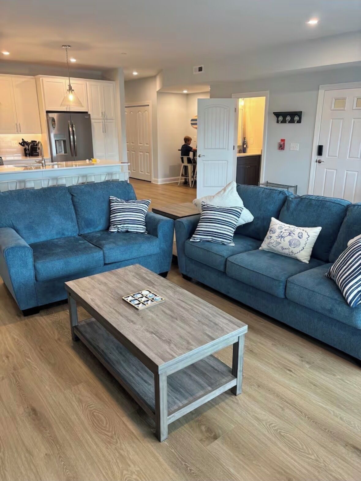 A modern living room with two blue sofas, a wooden coffee table, and a kitchen in the background.
