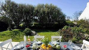 Une table à manger d’extérieur, avec des chaises blanches, des assiettes et un panier de fruits.
