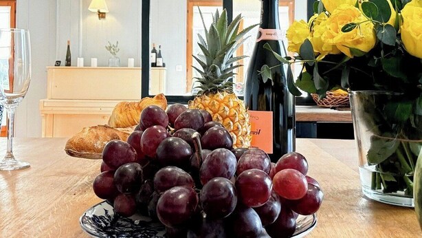 Une table à manger avec une assiette de raisins, une bouteille de vin, un ananas et un vase de fleurs jaunes.