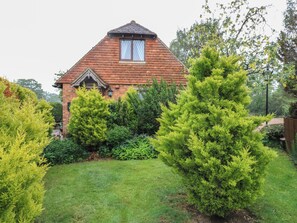 A brick house with a gabled roof, a small window, and a well-maintained garden with various shrubs and a lawn. - Barclay Cottage (Ashford)