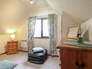 A bedroom with a window, a bedside table with a lamp, a dresser, a framed picture, and a small green glass container. - Barclay Cottage (Ashford)