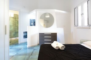 Ein Schlafzimmer mit einem schwarzen Bett, einem runden Spiegel, einer Kommode mit Schubladen und einem Fenster mit Jalousien.