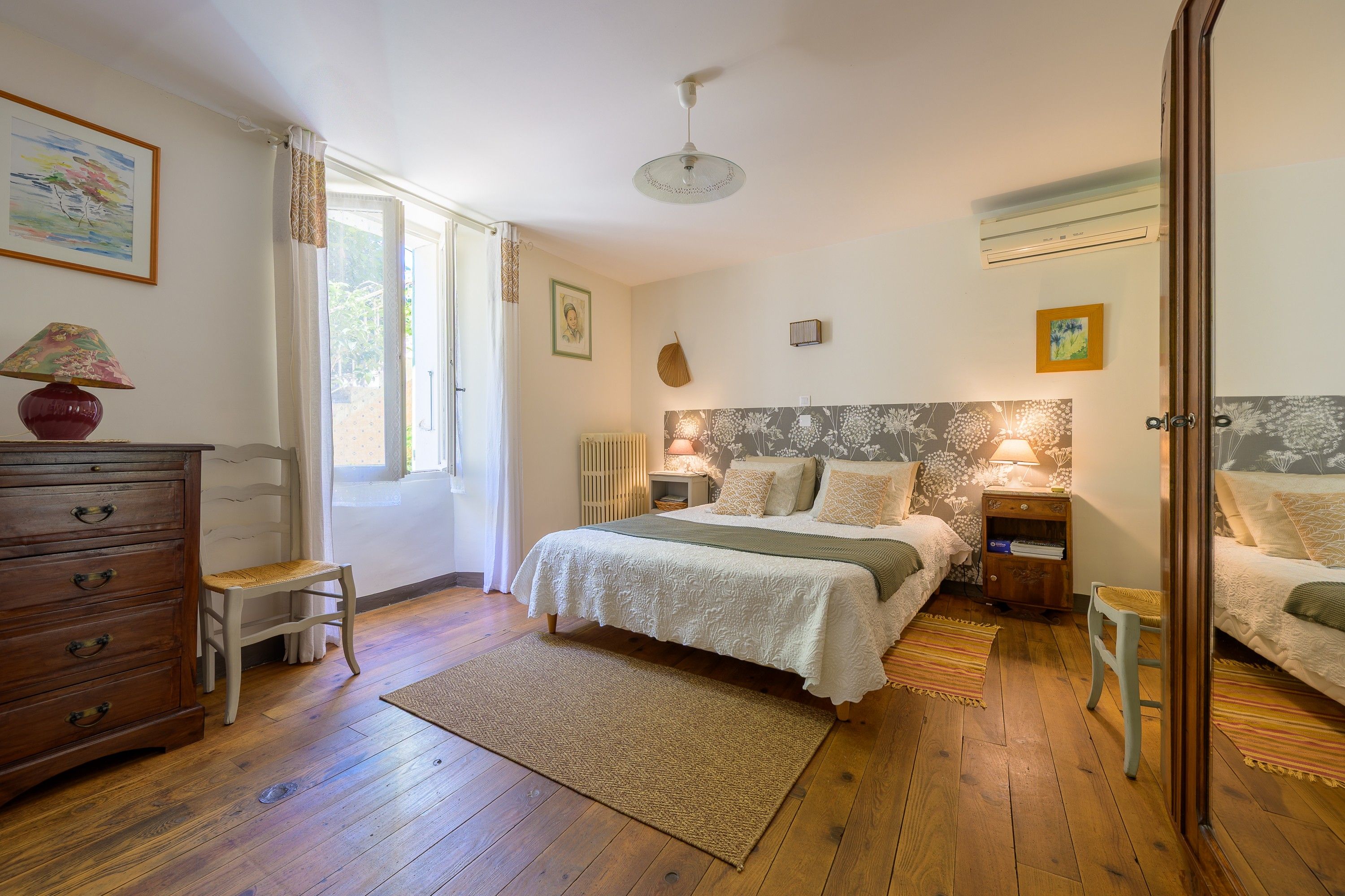 Une chambre avec un plancher en bois, un lit recouvert d’une couette blanche, une table de chevet avec une lampe, une commode et une fenêtre avec des rideaux.
