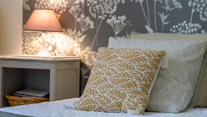 Un letto rifatto con cura, un cuscino decorato, una lampada da comodino e una carta da parati con motivi floreali.