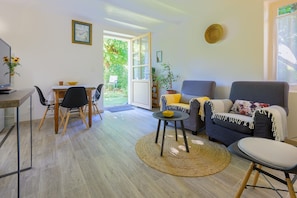 Een woonkamer met een bank, een eettafel met stoelen, een klein rond tafeltje met een schaal, en een groot raam met uitzicht op groen.