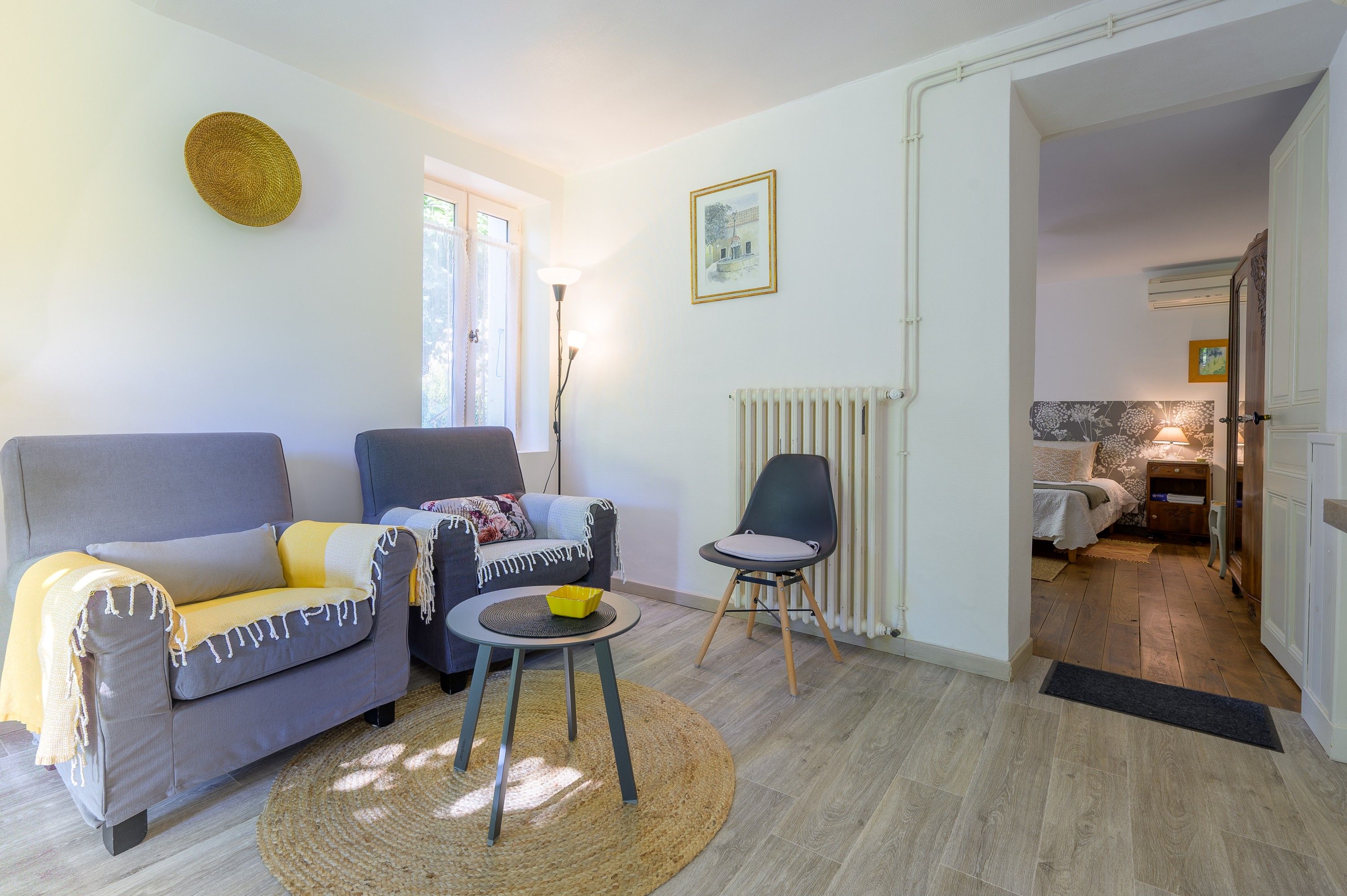 Un salon avec un canapé gris, un fauteuil plus petit, une petite table sur laquelle se trouve un bol jaune, et un radiateur.